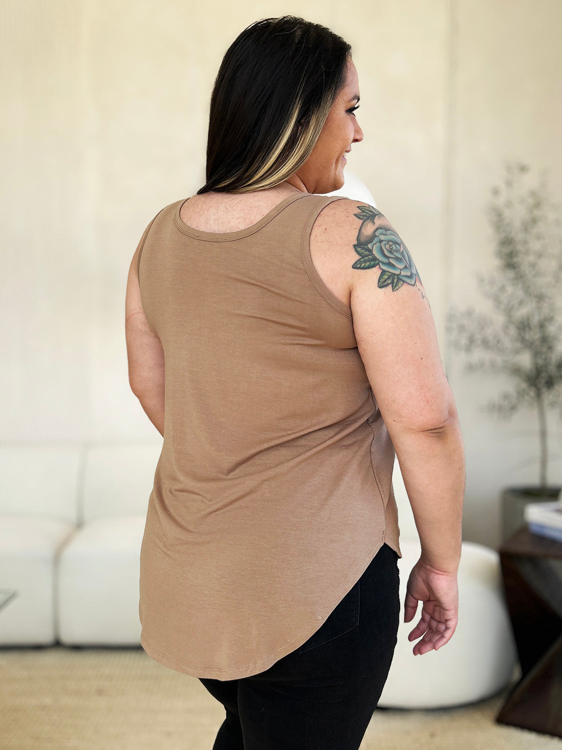 A woman wearing sunglasses, blue jeans, and the Basic Bae Full Size Round Neck Curved Hem Tank is standing in front of a concrete wall, holding her glasses with one hand and carrying a beige shoulder bag. Her basic style complements the slightly stretchy fabric of her outfit, ensuring comfort while maintaining chic simplicity.