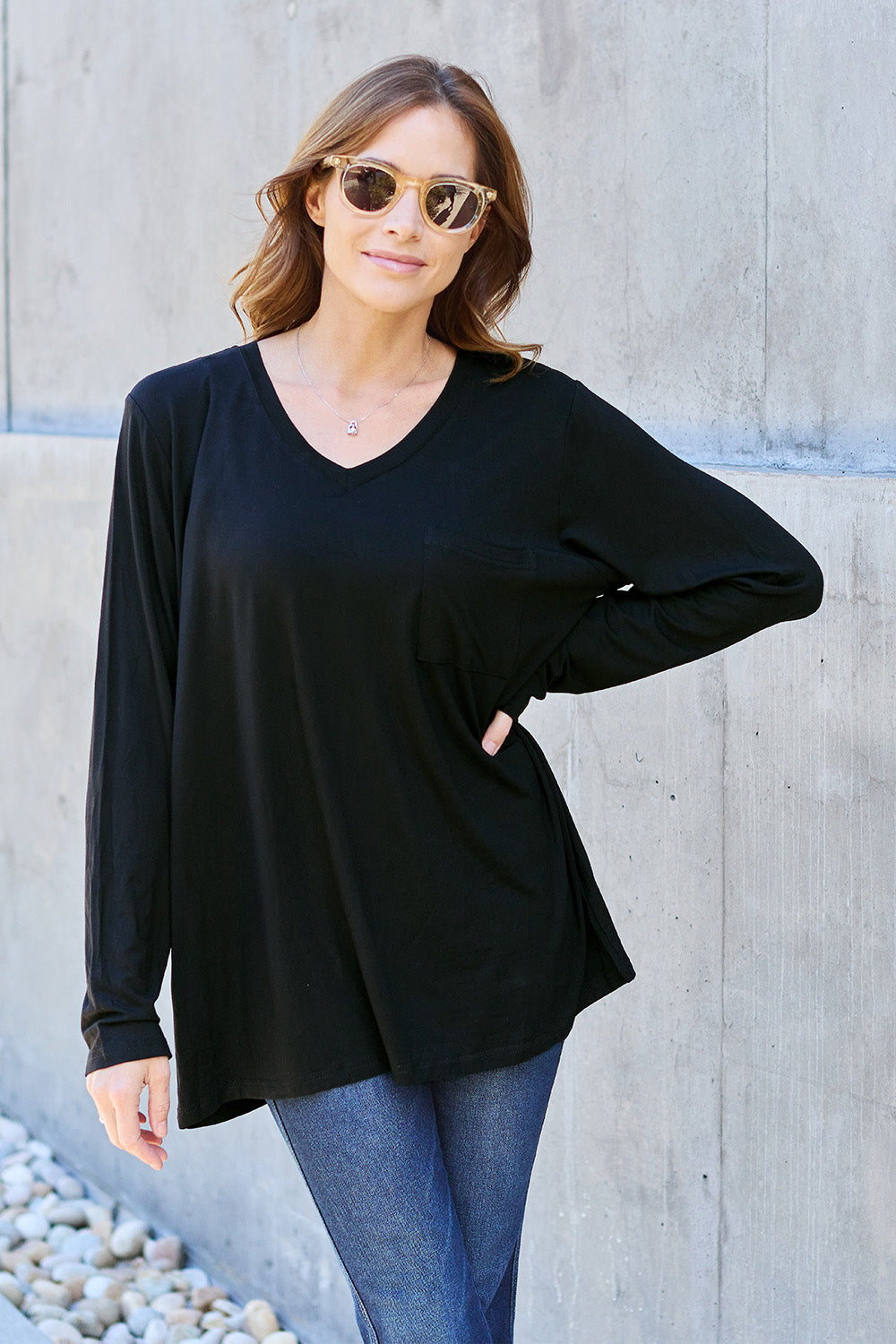 A woman with light brown hair stands against a concrete wall, wearing the Basic Bae Full Size V-Neck Long Sleeve Top in olive green and blue jeans, with a white backpack on her shoulder. Her outfit exudes basic style and is made from polyester, ensuring it’s both comfortable and suitable for machine wash cold. She is looking at the camera.