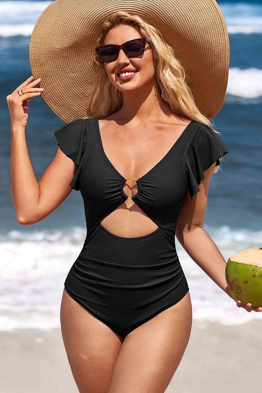 A woman in a Ruffle O-Ring Cutout One Piece Bathing Suit, large hat, and sunglasses stands on the beach holding a coconut.