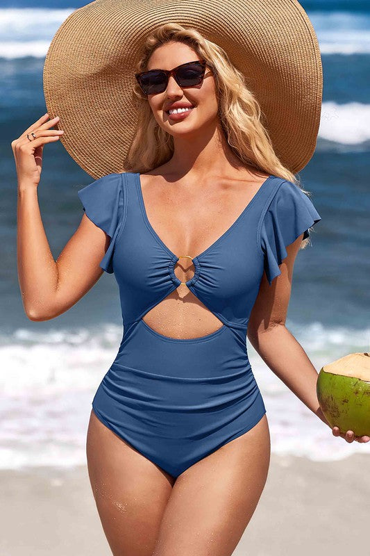 A woman in a Ruffle O-Ring Cutout One Piece Bathing Suit, large hat, and sunglasses stands on the beach holding a coconut.