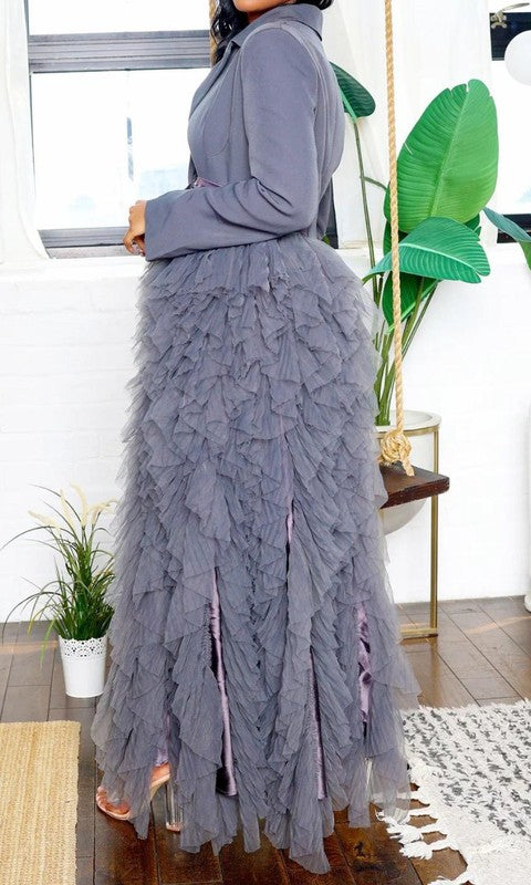 Person wearing the Noir Cascade Tuxedo Gown, which features structured shoulders, a deep V neckline, and a cascading ruffle skirt, paired with clear high-heeled shoes, exuding couture elegance.