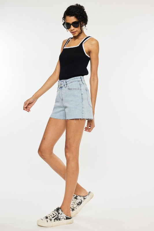 A person wearing a black top, High Rise Crossover WB Shorts in light stone wash denim, and black sunglasses, standing with crossed arms and a slight lean.