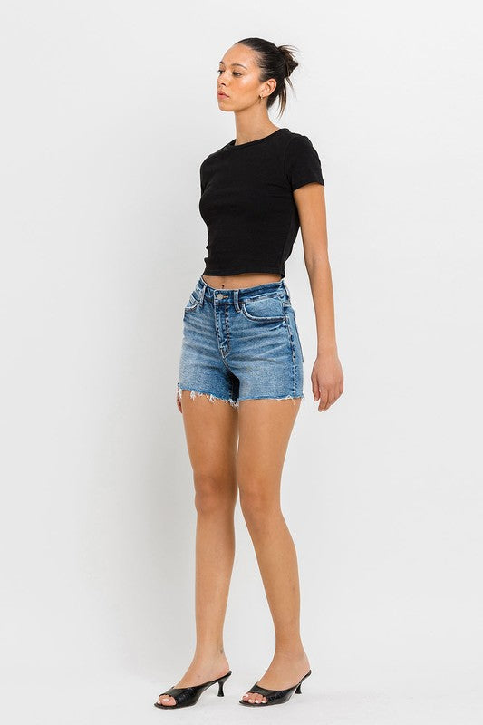 A person wearing a black top, High Rise Raw Hem Denim Shorts, and black sandals stands against a plain background.