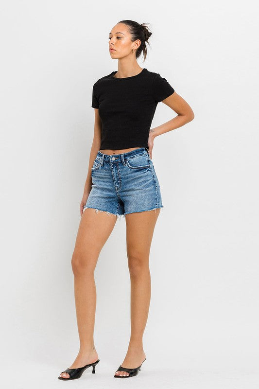 A person wearing a black top, High Rise Raw Hem Denim Shorts, and black sandals stands against a plain background.