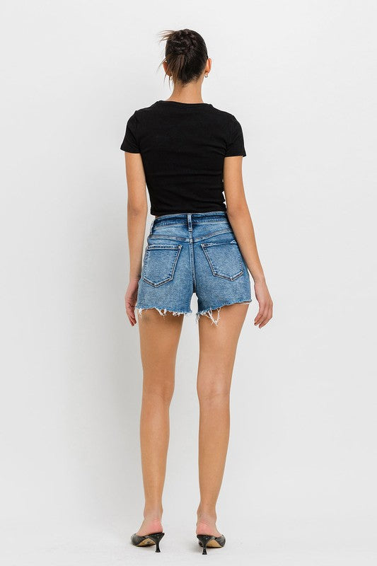 A person wearing a black top, High Rise Raw Hem Denim Shorts, and black sandals stands against a plain background.