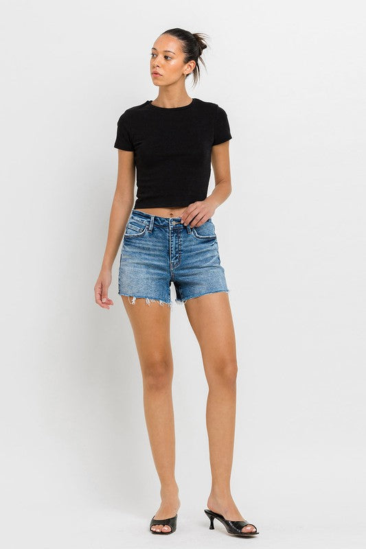 A person wearing a black top, High Rise Raw Hem Denim Shorts, and black sandals stands against a plain background.