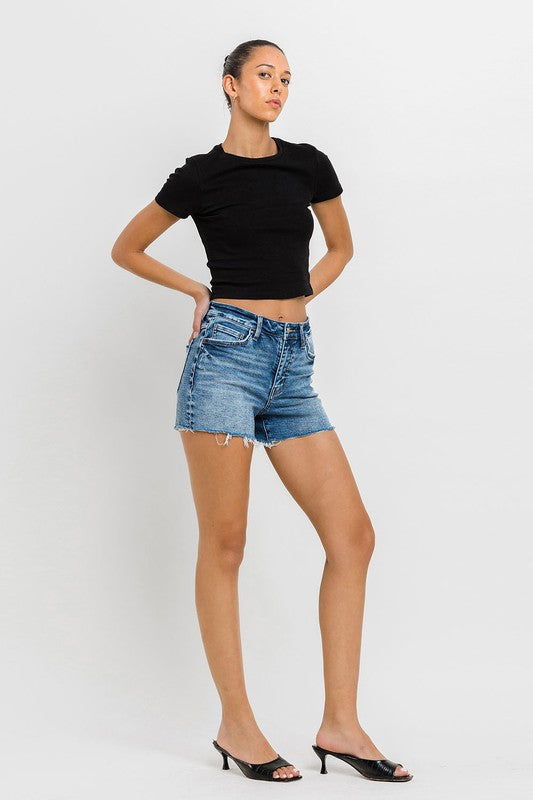 A person wearing a black top, High Rise Raw Hem Denim Shorts, and black sandals stands against a plain background.