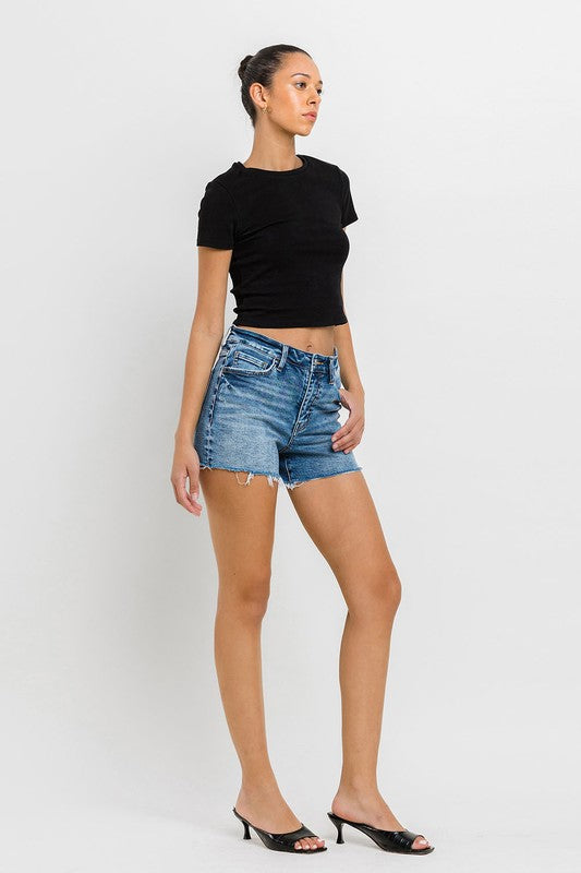 A person wearing a black top, High Rise Raw Hem Denim Shorts, and black sandals stands against a plain background.