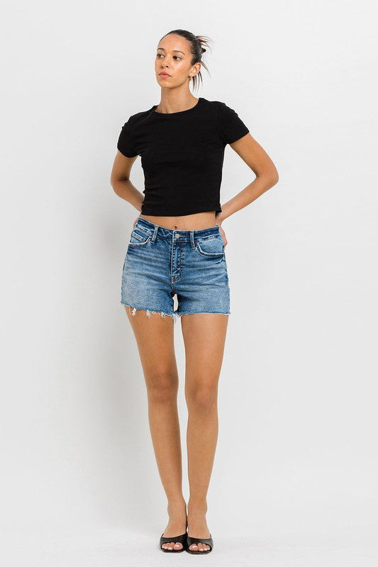 A person wearing a black top, High Rise Raw Hem Denim Shorts, and black sandals stands against a plain background.