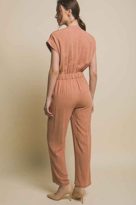 Wearing the V-Neck Pocketed Jumpsuit with buttons, a person pairs it with beige heels, standing casually against a plain backdrop, hands tucked in pockets.