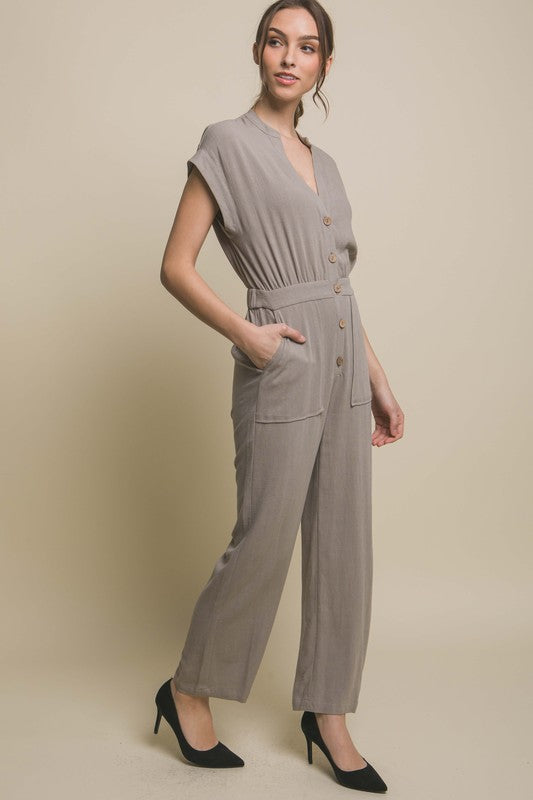 Wearing the V-Neck Pocketed Jumpsuit with buttons, a person pairs it with beige heels, standing casually against a plain backdrop, hands tucked in pockets.