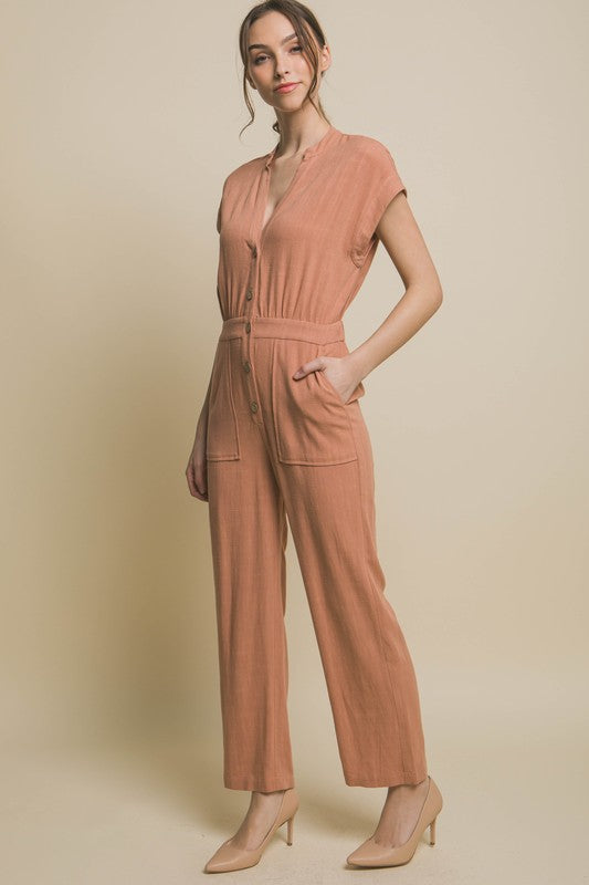 Wearing the V-Neck Pocketed Jumpsuit with buttons, a person pairs it with beige heels, standing casually against a plain backdrop, hands tucked in pockets.