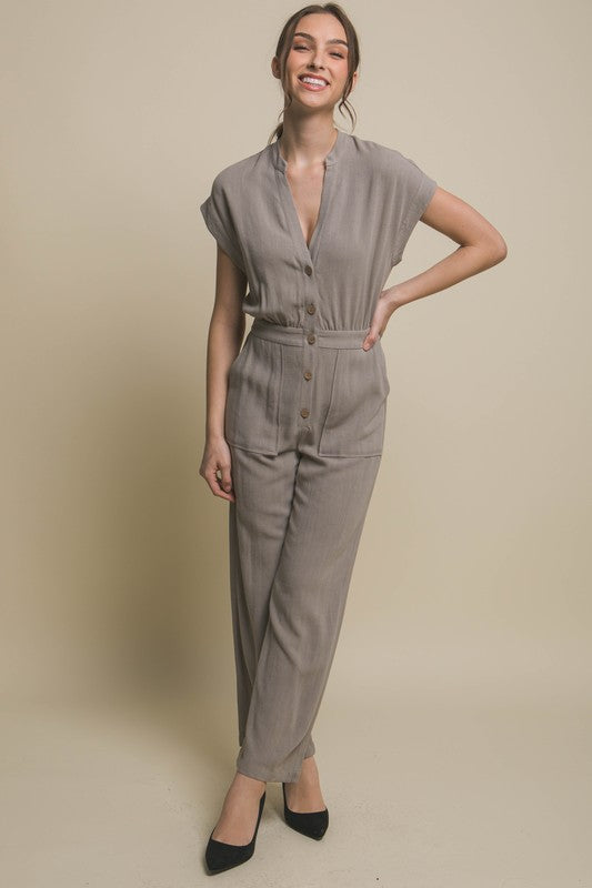 Wearing the V-Neck Pocketed Jumpsuit with buttons, a person pairs it with beige heels, standing casually against a plain backdrop, hands tucked in pockets.