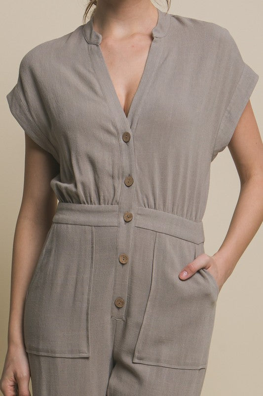 Wearing the V-Neck Pocketed Jumpsuit with buttons, a person pairs it with beige heels, standing casually against a plain backdrop, hands tucked in pockets.