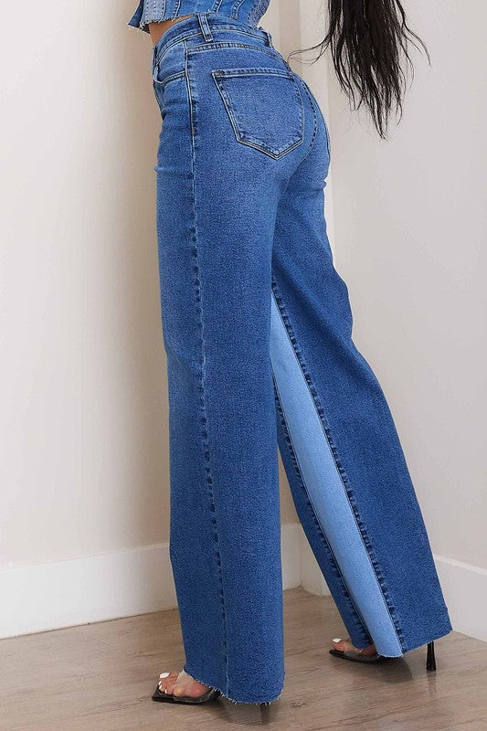 A woman, wearing a strapless denim top and High-Rise Color Block Wide Jeans, leans against a wall on a wooden floor. She has one hand in her pocket and the other resting on the wall.