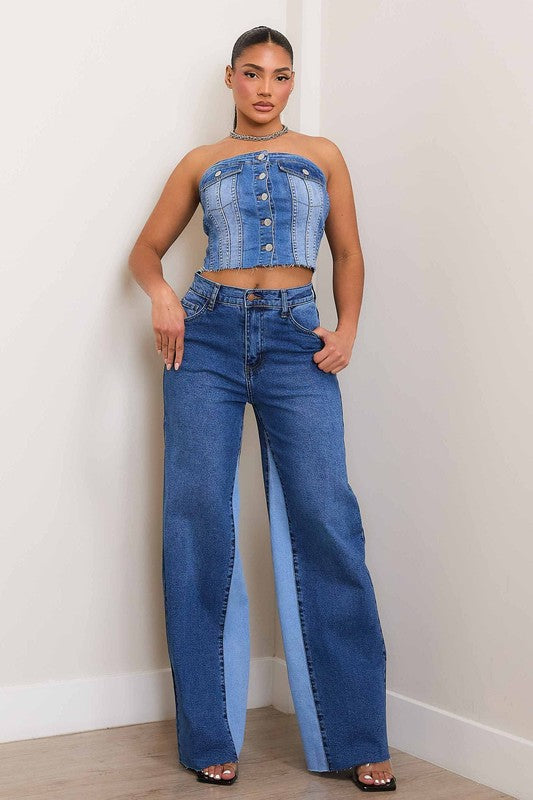 A woman, wearing a strapless denim top and High-Rise Color Block Wide Jeans, leans against a wall on a wooden floor. She has one hand in her pocket and the other resting on the wall.