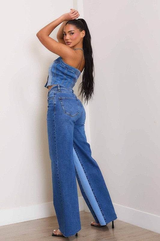 A woman, wearing a strapless denim top and High-Rise Color Block Wide Jeans, leans against a wall on a wooden floor. She has one hand in her pocket and the other resting on the wall.