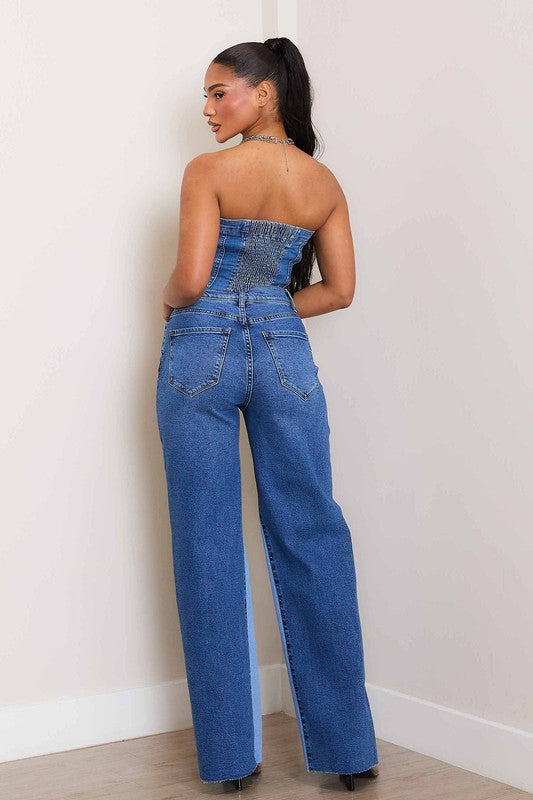 A woman, wearing a strapless denim top and High-Rise Color Block Wide Jeans, leans against a wall on a wooden floor. She has one hand in her pocket and the other resting on the wall.