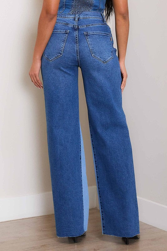 A woman, wearing a strapless denim top and High-Rise Color Block Wide Jeans, leans against a wall on a wooden floor. She has one hand in her pocket and the other resting on the wall.