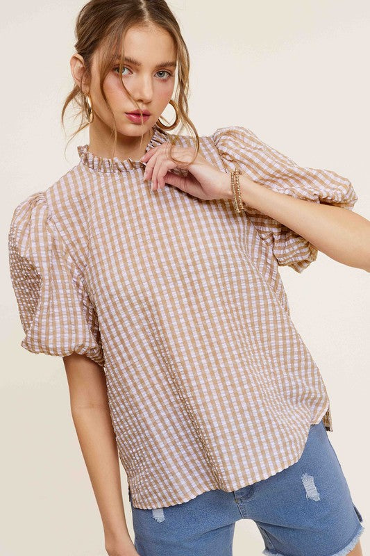 A person wearing a Gingham Check Print Puff Sleeve Top with a button-back closure sits on a chair, looking toward the camera.