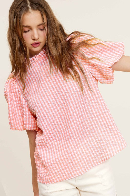 A person wearing a Gingham Check Print Puff Sleeve Top with a button-back closure sits on a chair, looking toward the camera.