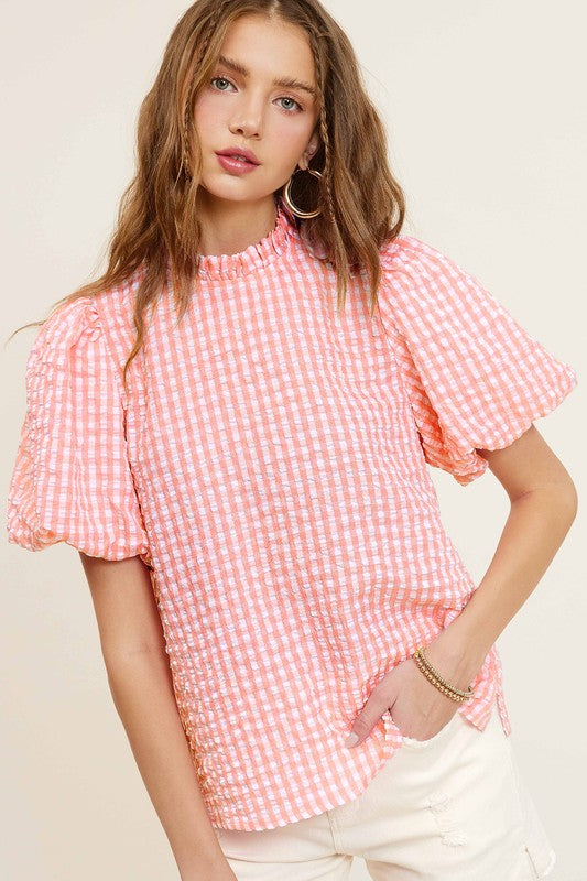 A person wearing a Gingham Check Print Puff Sleeve Top with a button-back closure sits on a chair, looking toward the camera.