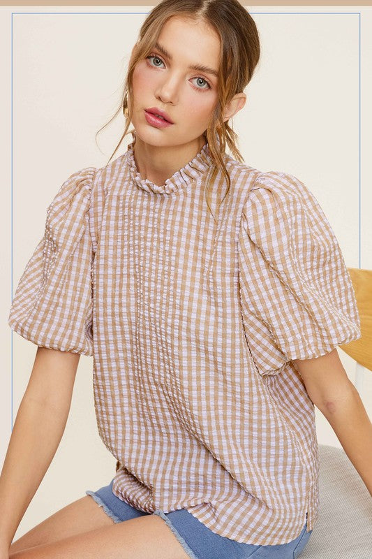 A person wearing a Gingham Check Print Puff Sleeve Top with a button-back closure sits on a chair, looking toward the camera.