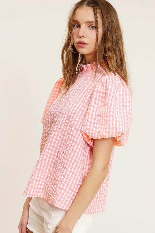 A person wearing a Gingham Check Print Puff Sleeve Top with a button-back closure sits on a chair, looking toward the camera.