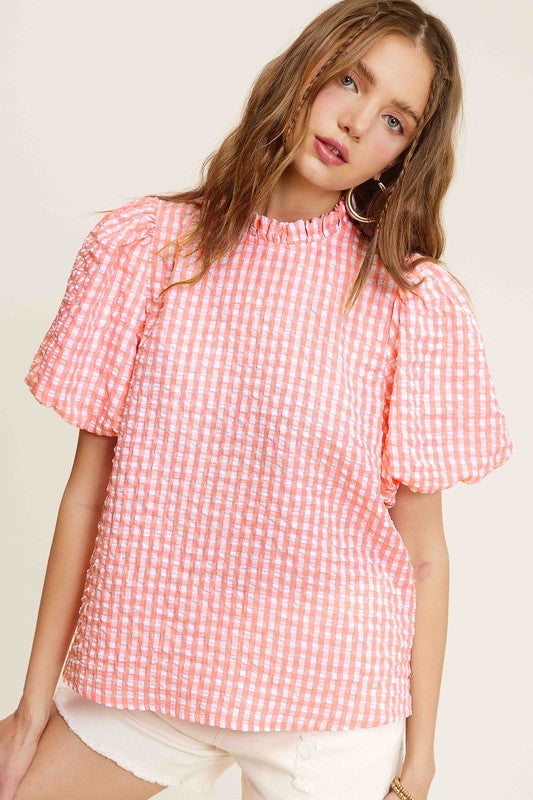 A person wearing a Gingham Check Print Puff Sleeve Top with a button-back closure sits on a chair, looking toward the camera.