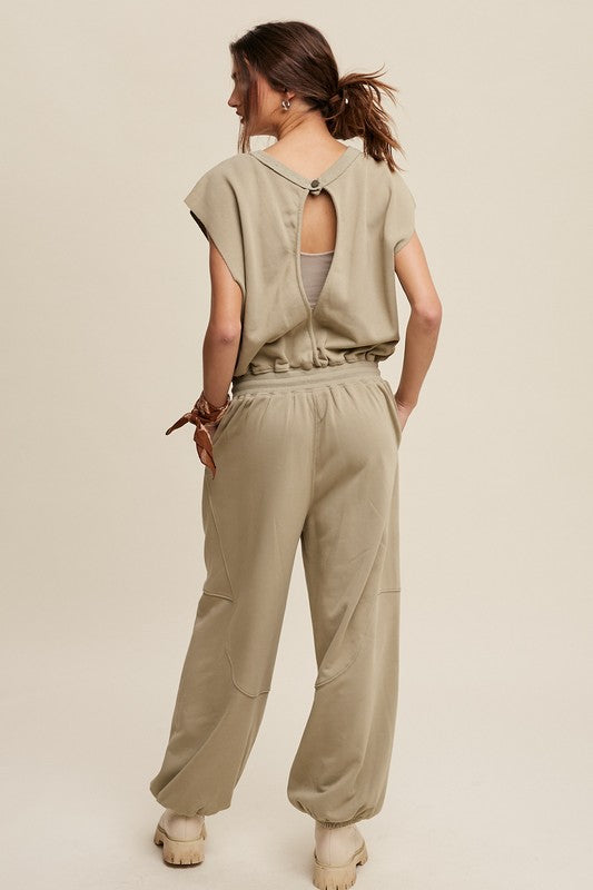 A person confidently walks against a plain background, dressed in an Athleisure French Terry Loose Jogger Jumpsuit and matching boots.