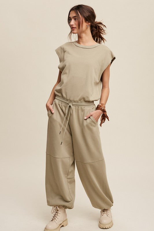 A person confidently walks against a plain background, dressed in an Athleisure French Terry Loose Jogger Jumpsuit and matching boots.