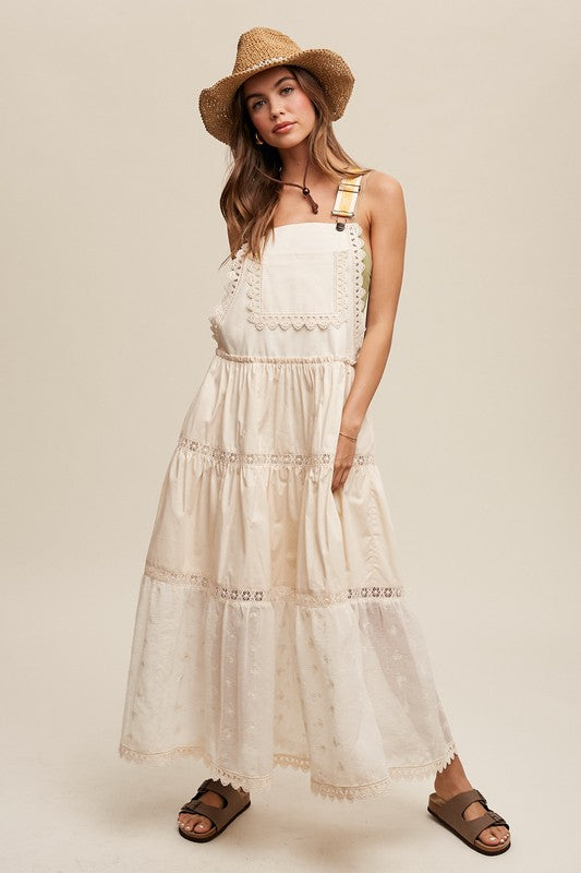 A person wearing the Laced and Tiered Romantic Overall Maxi Dress, complemented by a straw hat and brown sandals, stands gracefully against a plain background.