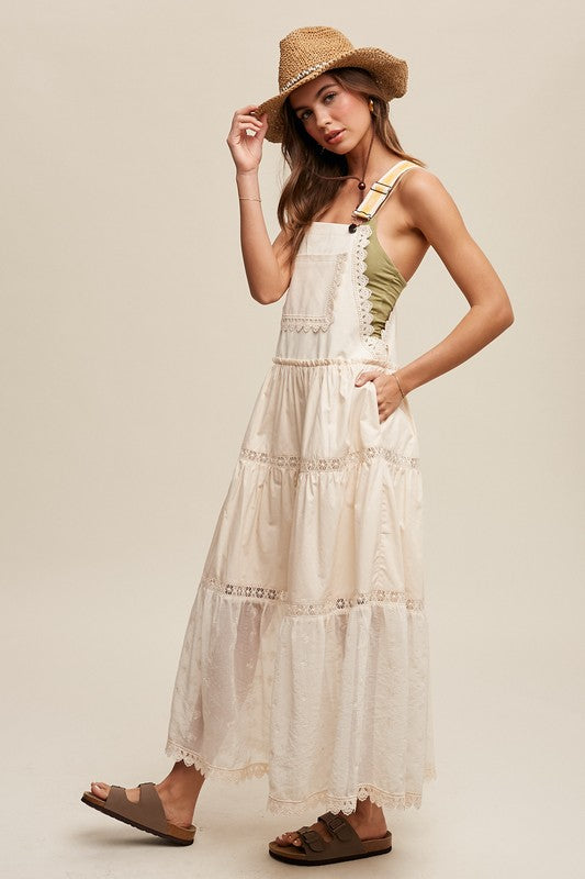 A person wearing the Laced and Tiered Romantic Overall Maxi Dress, complemented by a straw hat and brown sandals, stands gracefully against a plain background.