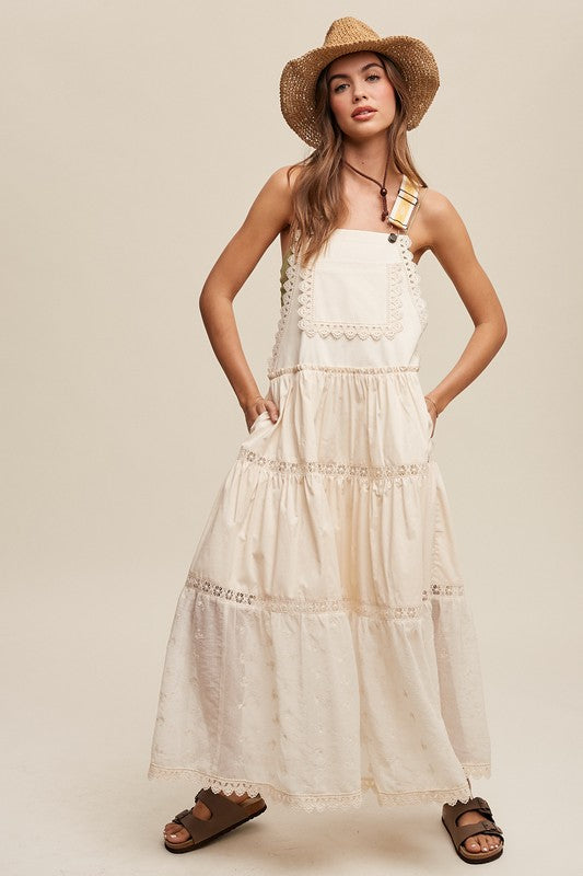 A person wearing the Laced and Tiered Romantic Overall Maxi Dress, complemented by a straw hat and brown sandals, stands gracefully against a plain background.