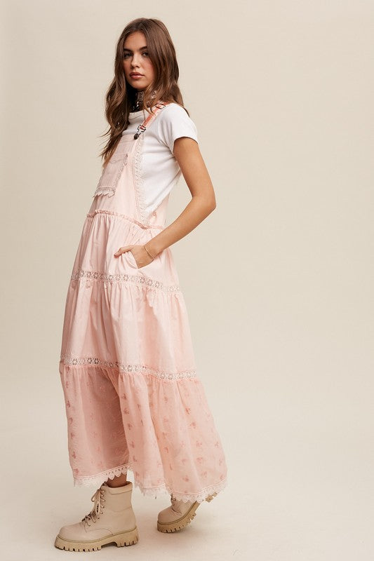 A person wearing the Laced and Tiered Romantic Overall Maxi Dress, complemented by a straw hat and brown sandals, stands gracefully against a plain background.