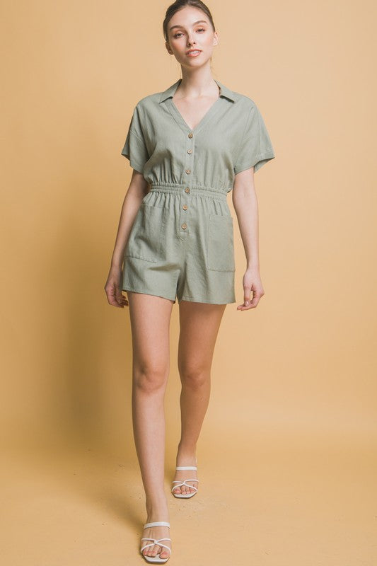 A woman stands against a beige background, wearing the Linen Button Down Romper in breathable linen fabric. The short-sleeved, green romper features a cinched waist and pockets, ideal for casual comfort.