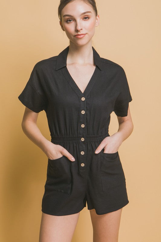 A woman stands against a beige background, wearing the Linen Button Down Romper in breathable linen fabric. The short-sleeved, green romper features a cinched waist and pockets, ideal for casual comfort.