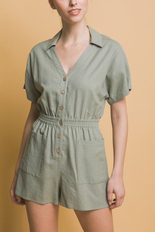 A woman stands against a beige background, wearing the Linen Button Down Romper in breathable linen fabric. The short-sleeved, green romper features a cinched waist and pockets, ideal for casual comfort.