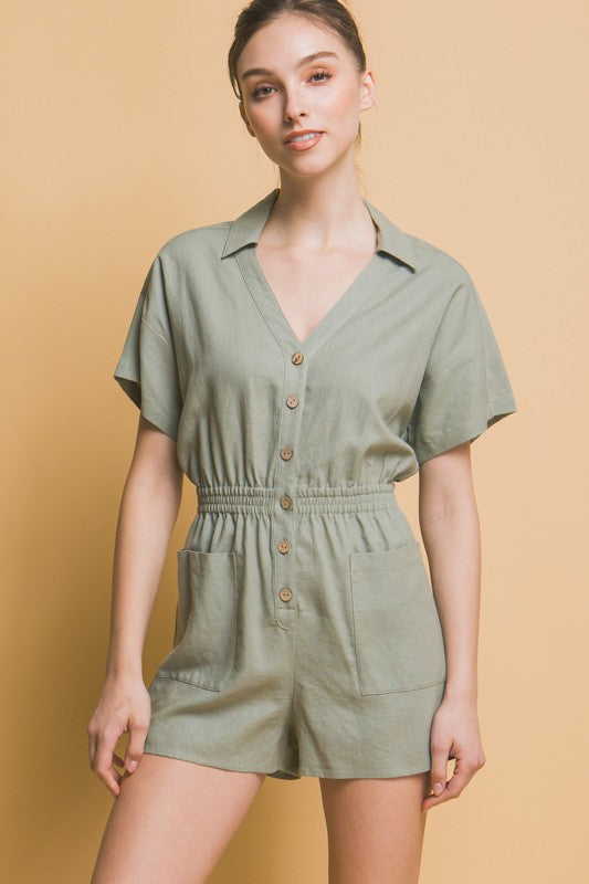 A woman stands against a beige background, wearing the Linen Button Down Romper in breathable linen fabric. The short-sleeved, green romper features a cinched waist and pockets, ideal for casual comfort.