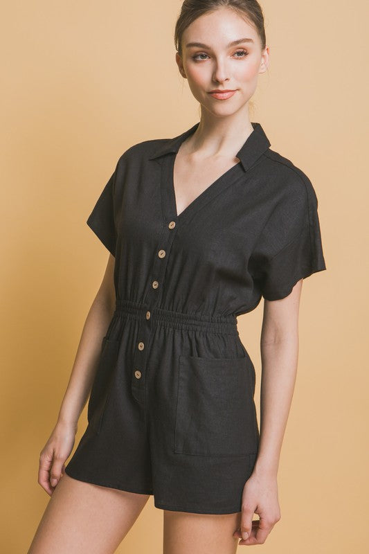 A woman stands against a beige background, wearing the Linen Button Down Romper in breathable linen fabric. The short-sleeved, green romper features a cinched waist and pockets, ideal for casual comfort.