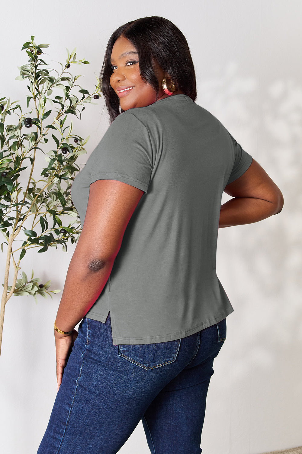 A person with wavy hair, wearing the Basic Bae Full Size Round Neck Short Sleeve T-Shirt in bright pink and beige pants, stands near a plant, smiling and looking to the side. The outfit embodies a basic style and offers slightly stretchy comfort, ideal for those who value effortless elegance.