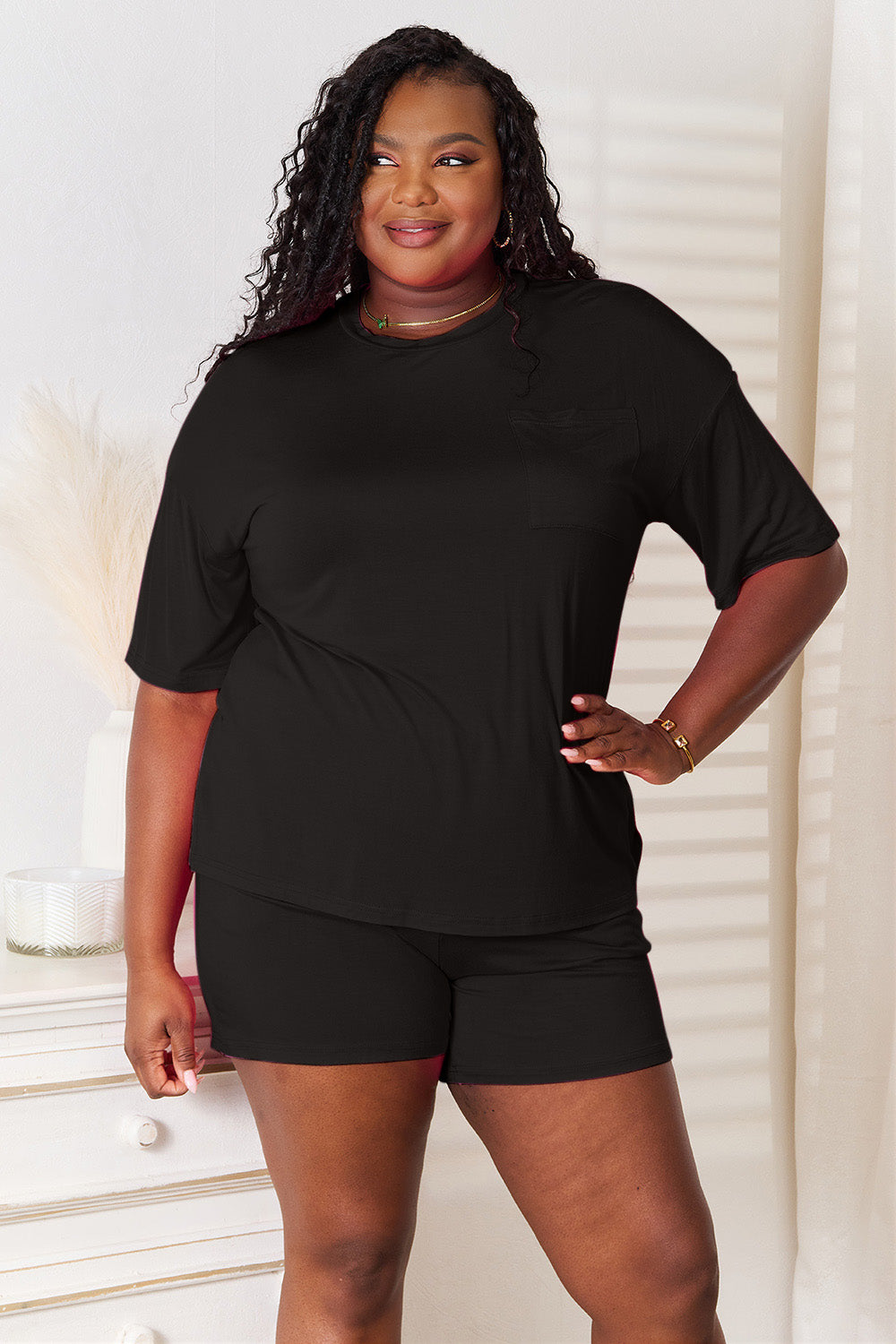 A woman is standing in a brightly lit room, wearing the Basic Bae Full Size Soft Rayon Half Sleeve Top and Shorts Set in a relaxed, stretchy turquoise fabric. She has one hand placed in her pocket and is smiling while looking slightly to the side.