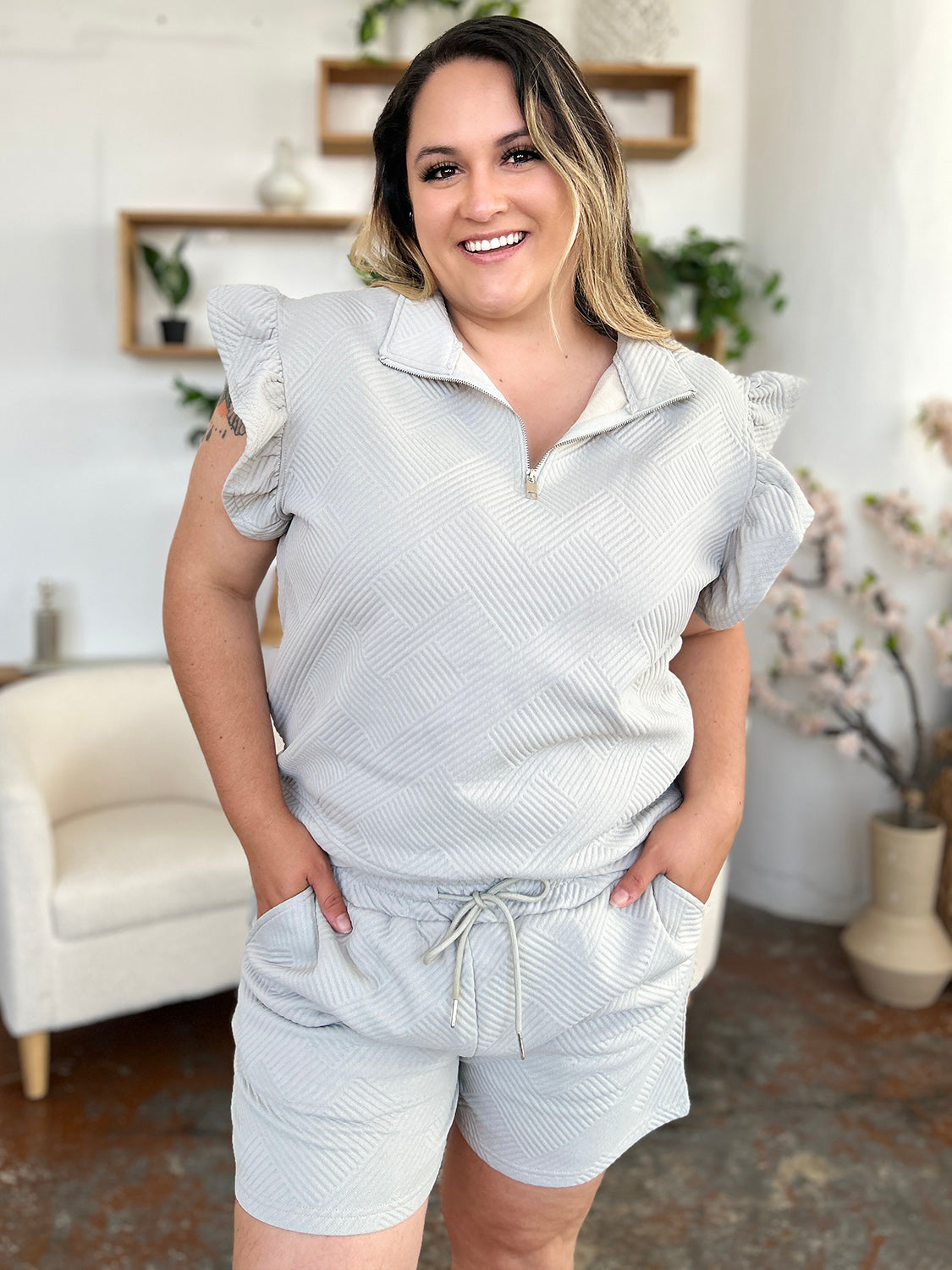 Against a plain background, a woman with blonde hair poses confidently with one hand in her pocket, highlighting the chic style of her Double Take Full Size Texture Flounce Sleeve Top and Drawstring Shorts Set.