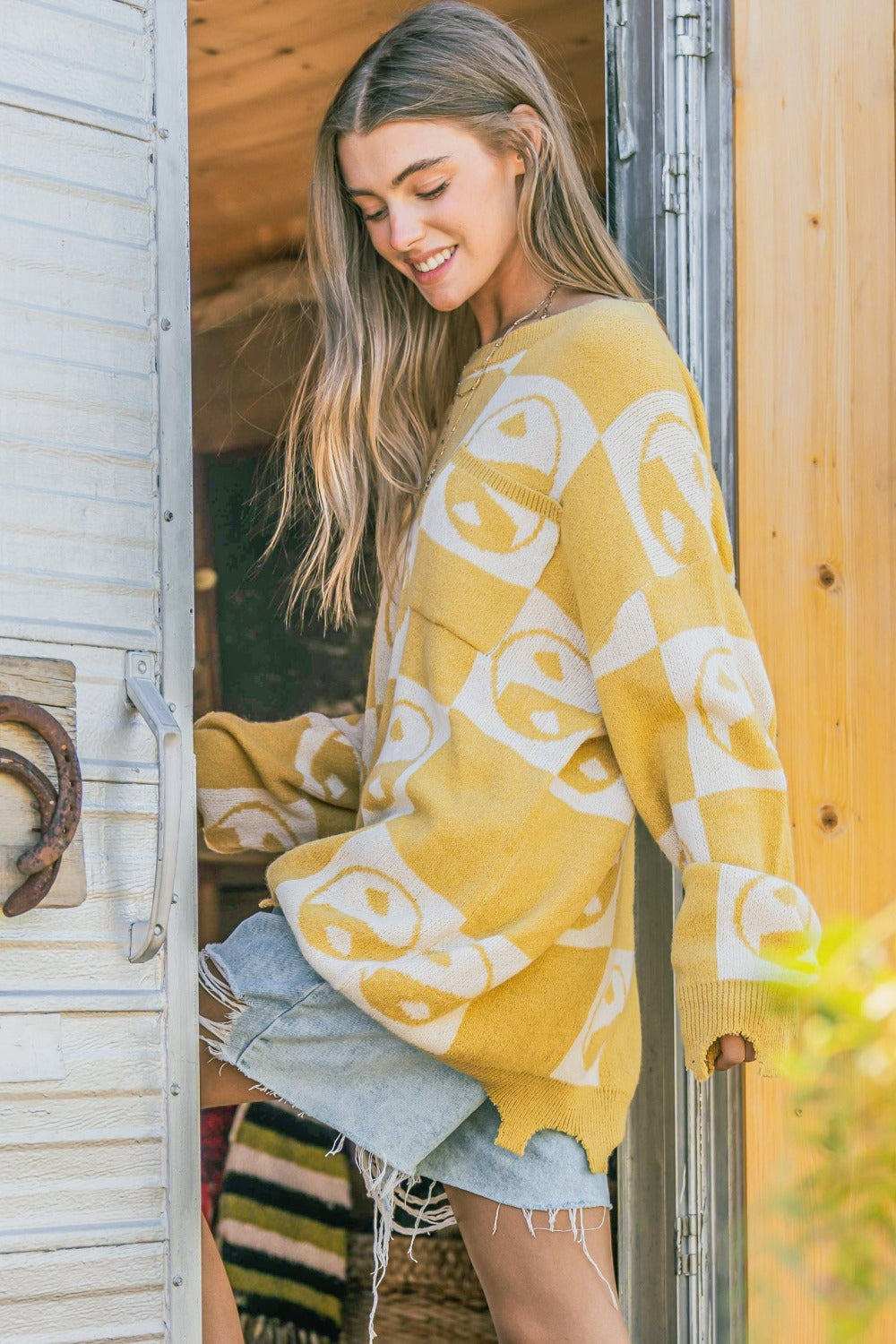 A woman wearing the And The Why Checkerboard Sweater with a Yin Yang pattern and distressed denim shorts stands in a doorway.