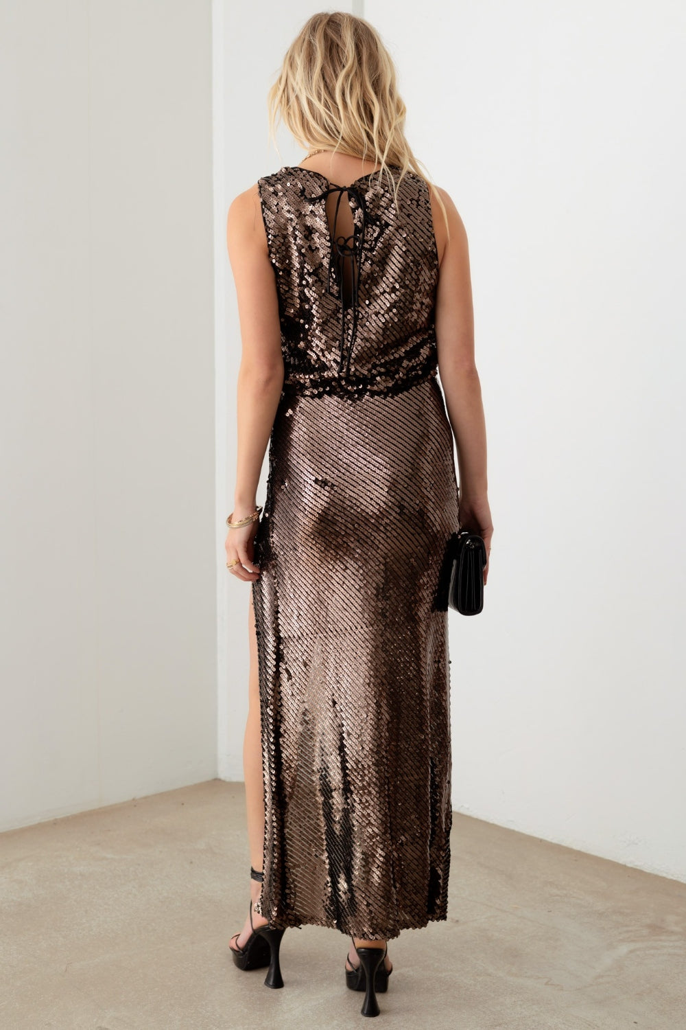 Woman in a Mustard Seed Metallic Shiny Sequins Back Ribbon Bubble Top, holding a small black clutch, standing indoors against a plain white background.