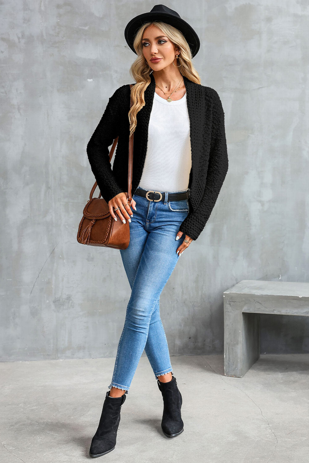 A woman in an Apricot Popcorn Knit Open Front Cardigan and jeans stands against a brick wall, looking to the side with a slight smile.