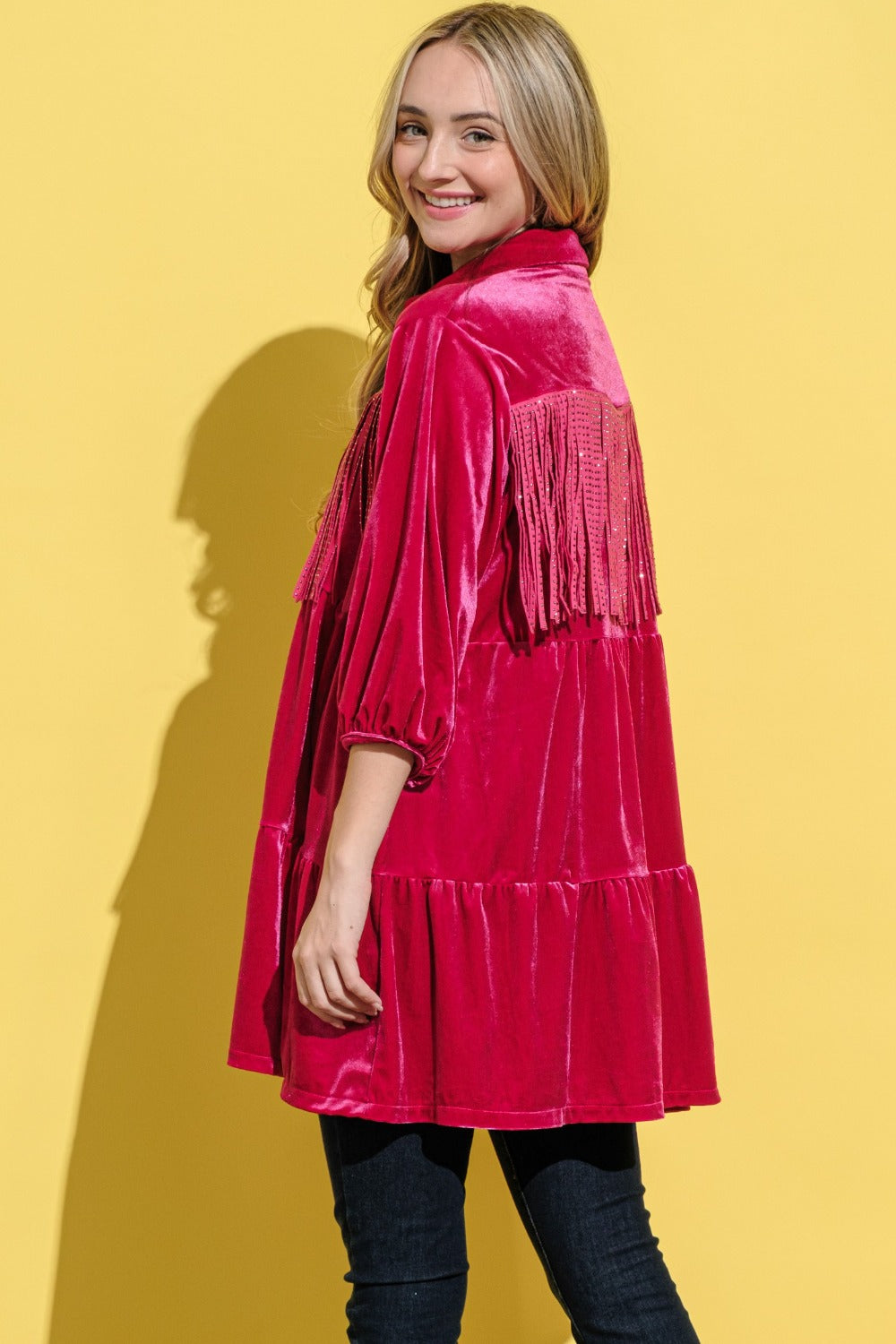 A woman stands against a bright yellow background, smiling and wearing an elegant statement fringed, bright pink And The Why Fringe Detailed Velvet Shirt Dress.