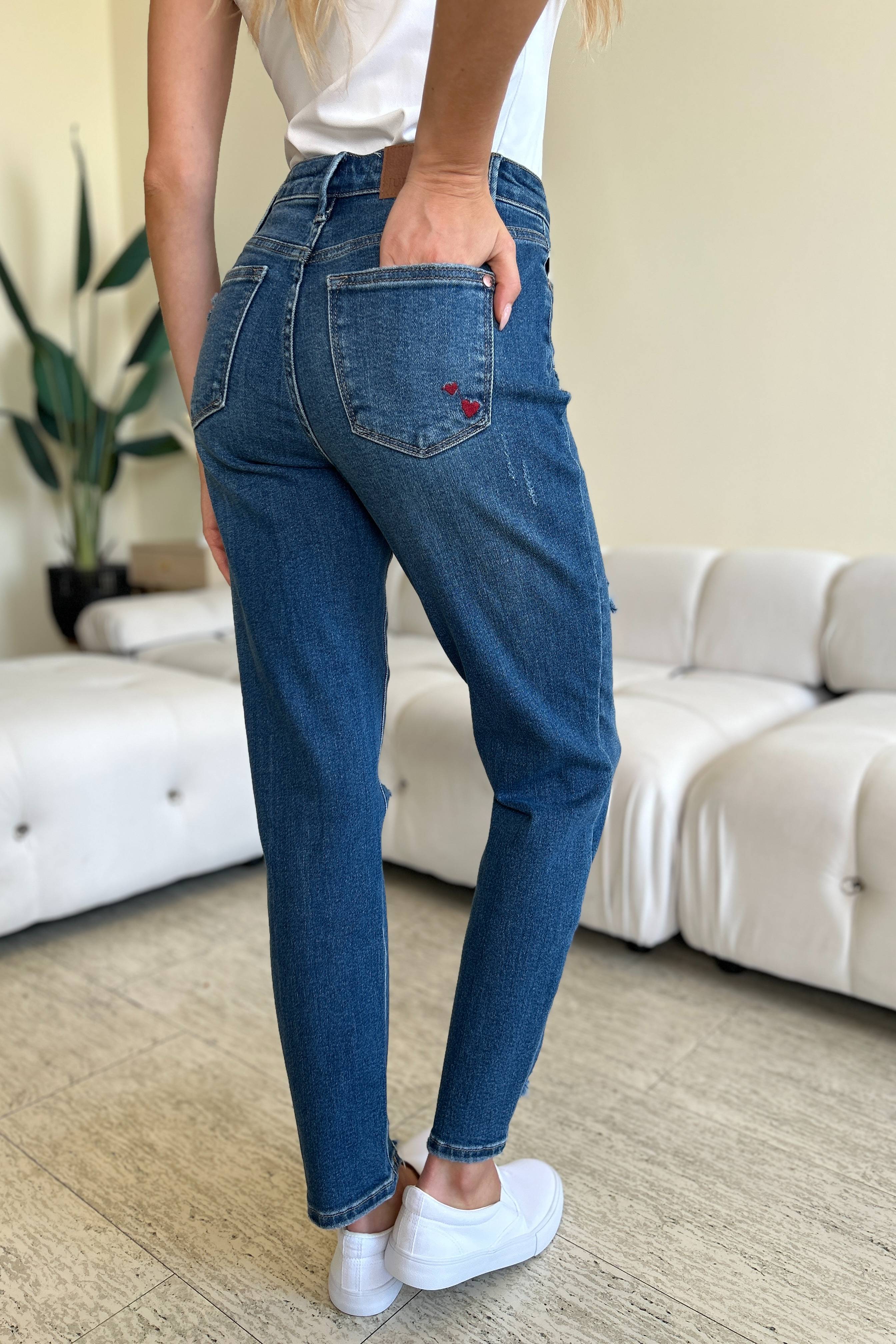 Two women stand side by side in a living room, both wearing stylish jeans and tank tops. One woman is pointing at the distinctive back pocket of her Judy Blue Full Size Queen Of Hearts Coin Pocket BF Jeans, while the other smiles.