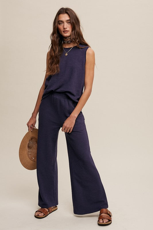 A person stands against a plain background wearing the navy blue Soft Knit Tank and Sweat Pant Set. They are holding a brown hat and appear to be wearing sandals. Their hand is in one pocket, epitomizing comfortable casual wear.