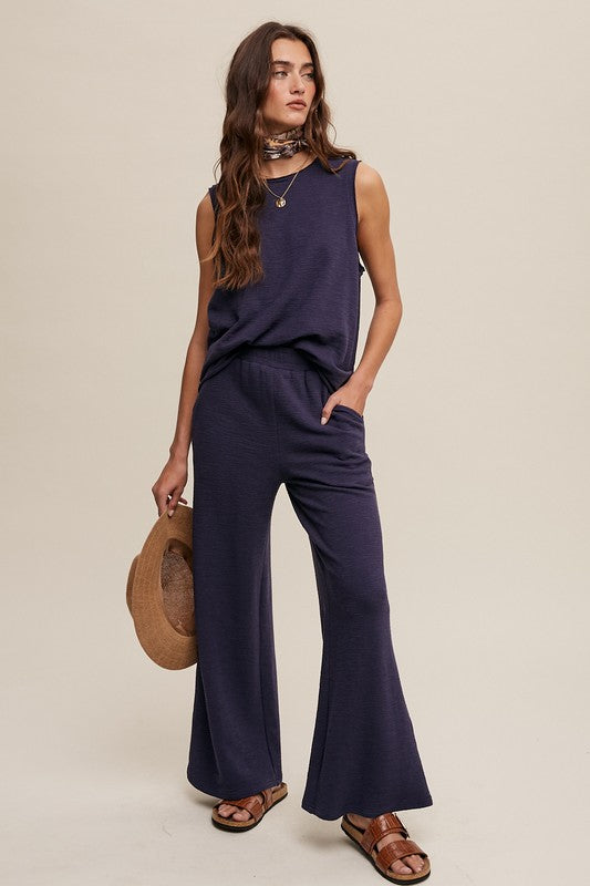 A person stands against a plain background wearing the navy blue Soft Knit Tank and Sweat Pant Set. They are holding a brown hat and appear to be wearing sandals. Their hand is in one pocket, epitomizing comfortable casual wear.